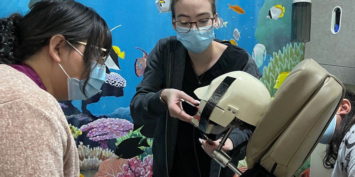 Two masked women adjusting a dental mannequin head in a colorful aquatic-themed room.